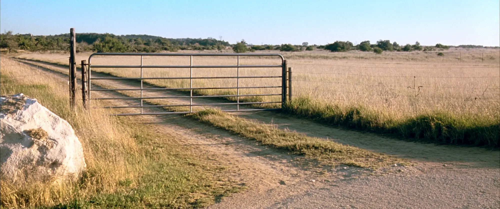 A closed farm gate on a dirt track leading into open Texas scrubland at late afternoon.